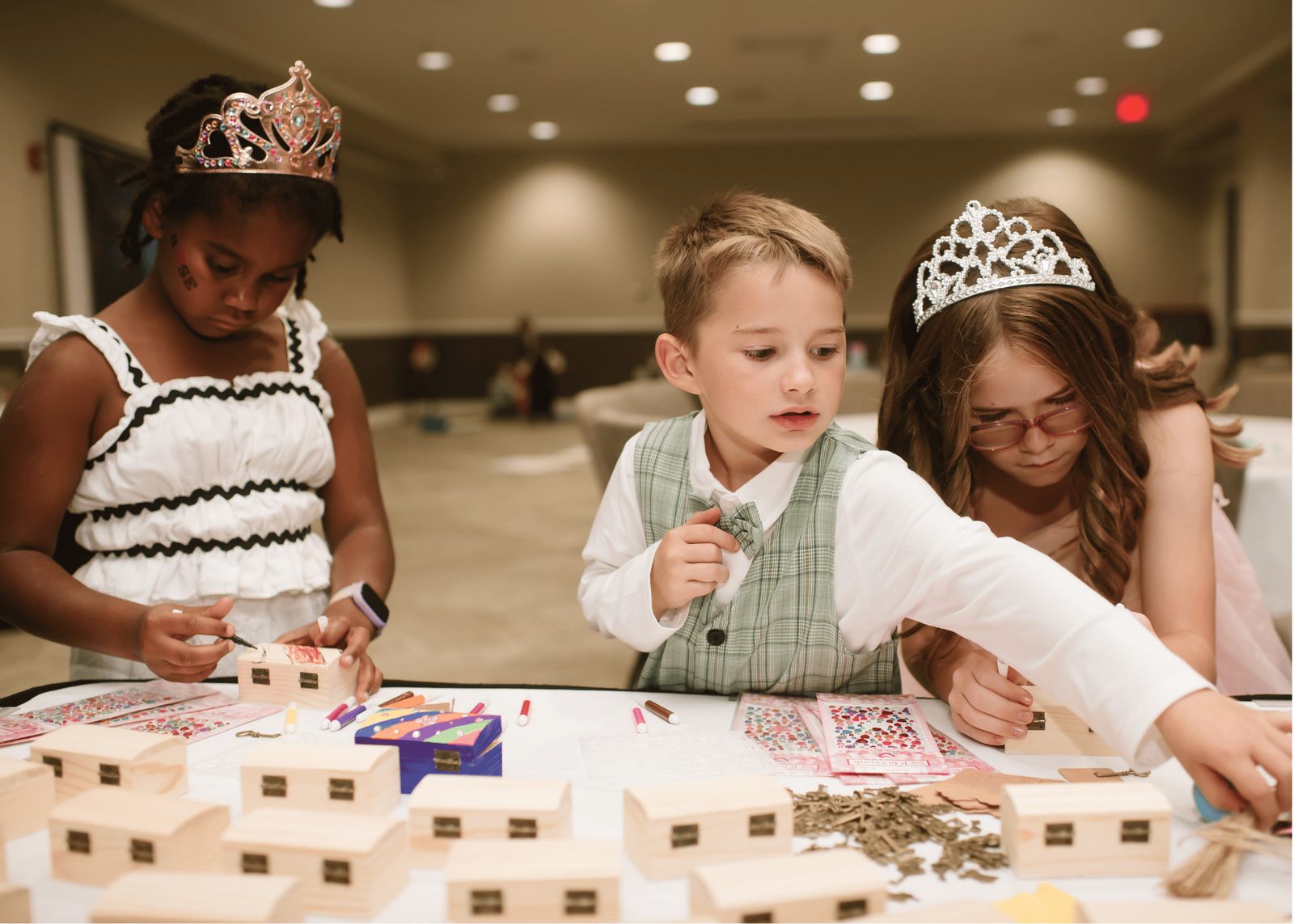 Children wearing tiaras decorate wooden treasure boxes at a craft table.